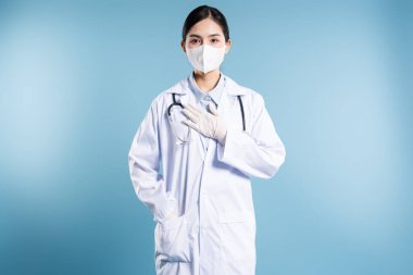 Beyaz önlük maskeli ve eldivenli bir doktorun ön çekimi mavi arka plan steteskopuna karşı görünür.