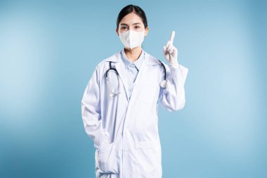 Maske eldivenli ve laboratuvar önlüklü bir doktor açık mavi bir stüdyo arkaplanını işaret ediyor.