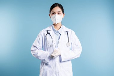 Laboratuvar önlüğü ve maskesiyle bir stüdyo ortamında duran sağlık görevlisi. Stetoskobu var.