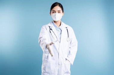 Kadın doktor yüz maskesi ve steteskoplu laboratuvar önlüğü giyerken mavi arka planda el sıkışır.