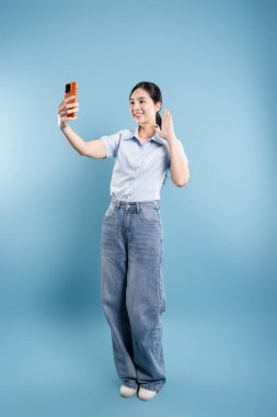 Turuncu mobil cihazlı gülümseyen kadın el sallarken açık mavi bir arkaplanda selfie çekiyor.