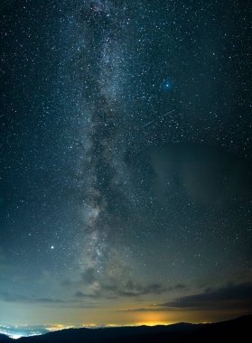 Milky way seen seen from the mountains, rising above urban city lights in a summer night. Astrophotography with stars and constellations. 