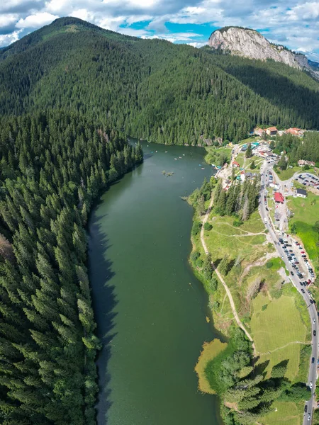 Aerial panorama of Red Lake and an asphalt road winding along its shores. The lake is surrounded by dense coniferous forests. A tourist station stands on the lake\'s shores.