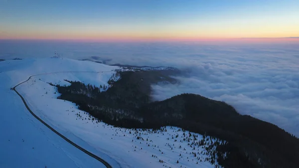 Aerial drone panorama of a winter sunset over Parang Mountains. A sea of clouds is covering the dense pine forests underneath. Everything is frozen.