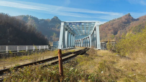 An iron train bridge over the Olt river. The railroad winds along the river's valley through rocky hills. Sunny day, the nearby forests are reflecting into the shallow water. Carpathia, Romania.