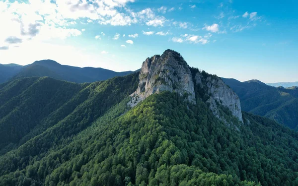 An eroded cliff pointing out of a beech forest on top of a mountain peak. The wild beech forest surrounding it are all green. Sunset time, sunlight shines through the clouds. Carpathia, Romania.