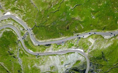 Dağlık çayırlarda dönen bir dağ yolunun üstündeki hava manzarası. Asfalt yol dağdan geçen arabalar tarafından geçiliyor. Yol boyunca bir dere akar. Carpathia, Romanya.