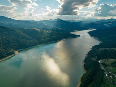 Izvorul Muntelui Gölü üzerindeki hava aracı panoraması. Güneş batmak üzere. Mavi sular yakındaki turistlerin tatil beldelerinin kıyılarını yıkıyor. Ceahlau Dağları, Karpatya.