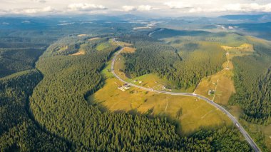 Bir dağ geçidinin üstündeki hava aracı manzarası üzerinde kozalaklı ormanlar olan sakin bir çayırı geçiyor. Ufkun altındaki dağ zirveleri boyunca asfalt bir yol esiyor. Gün batımı zamanı.
