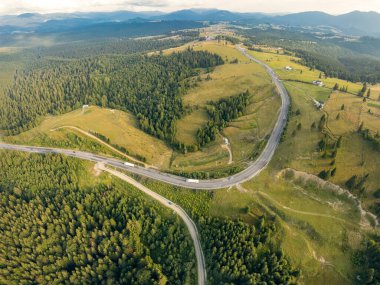 Bir dağ geçidinin üstündeki hava aracı manzarası üzerinde kozalaklı ormanlar olan sakin bir çayırı geçiyor. Dağ tepeleri boyunca asfalt bir yol esiyor. Üzerinde bir sürü araç var. Gün batımı zamanı.