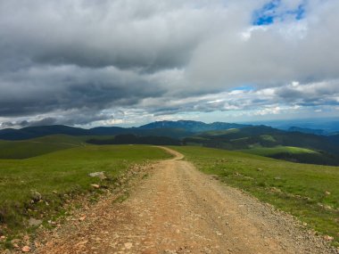 Dağlık tepelerin ve dağlık tepelerin yakınındaki dağlık çayırlarda dolanan toprak bir yol. Bulutlar gölgelerini dağ geçidine bırakıyor. Carpathia, Romanya.