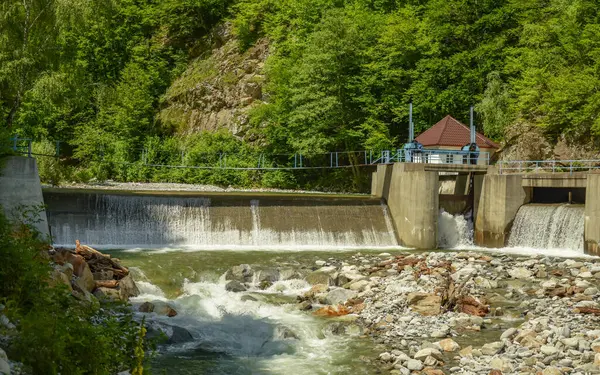 stock image A small power plant with its concrete dam built on a mountain river. The water is overflowing the built structure. An old footbridge pontoon crosses the small lake. Green energy, Carpathia, Romania.