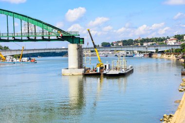 Tarihi Sava Demiryolu Köprüsü sökülüyor; ağır makineler, nehir yansımaları ve eski ve Yeni Belgrad kontrastı.