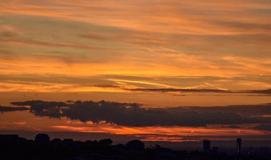 Batan güneş, evlerin ve binaların siluetleri çarpıcı bir şehir manzarası oluştururken turuncu tonlarda gökyüzünü aydınlatıyor..