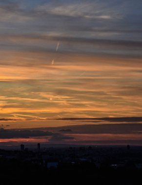 Aydınlatılmış bulutlar ve sıcak renk geçişleriyle dramatik akşam batımı, siluetli şehir manzarası yukarıdaki parlayan gökyüzünü yükseltiyor.