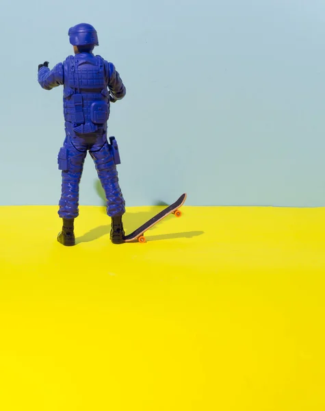 The policeman is holding a skateboard with his foot. The background is very yellow and pastel blue. The conceptual setting reflects the contrast between cheerful relaxation and severity.