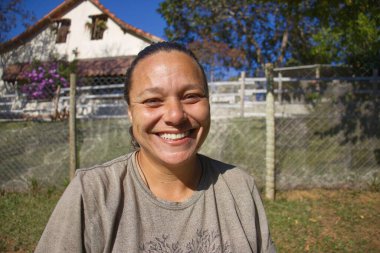 Gülümseyen bir kadın açık havada, yemyeşil bir arka planda oturur. Gri bir tişört giyiyor ve neşe saçıyor. Arka planda, bir ev ve yeşil bitki örtüsü sahneyi tamamlıyor..