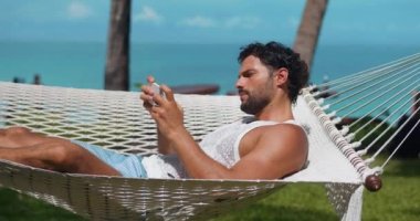 Bronzlaşmış tenli, akıllı telefondan online oyunlar oynayan yaz tatilinde hamakta uzanan hoş bir adam.