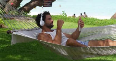Bronz tenli, kulaklıklı, akıllı telefonuyla radyo dalgalarını ayarlayan, hamakta dinlenen yakışıklı bir turist.
