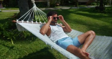 Tropik bir kaçamak. Egzotik geri çekilme. Güneş banyosu. Yakışıklı turist güneş gözlüğü takıyor ve plaj hamağında dinleniyor.