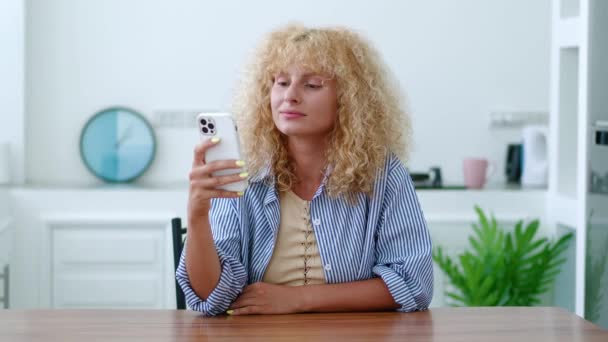 スタイリッシュなカールを持つ美しい赤髪の女性 携帯電話を使用して 夢のようなニュースを読んで 家のインテリアのテーブルに座って — ストック動画