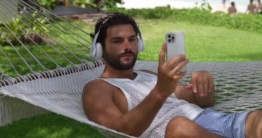 Kulaklıklı yakışıklı bir turist, hamakta dinleniyor, akıllı telefon kullanıyor, müzik parçalarını ayarlıyor, haber yayını yapıyor.