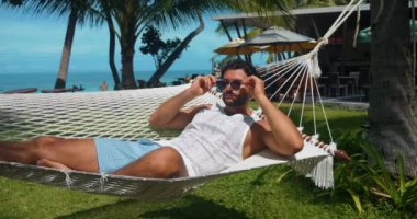 Kafkasyalı adam güneş gözlüğü takar, plaj kıyısındaki tatil beldesinde iki palmiye ağacı arasında kamp yaparken ürperir.