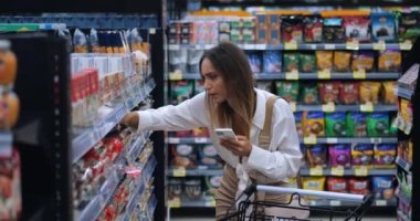 Genç, kendine güvenen Avrupalı kadın tüketici, akıllı telefon kullanan alıcı, gıda süpermarketinden alışveriş yaparken