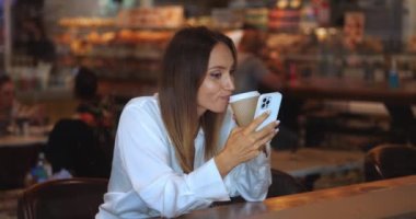Bir kafede kahve içen, akıllı telefonunu kullanan, haber kaynaklarını kaydıran, sosyal medya içeriğini kontrol eden çekici genç bir kadın.