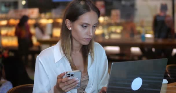 头晕的女人在电话中仔细检查信息 用笔记本电脑检查 在咖啡店的内部远程工作 — 图库视频影像