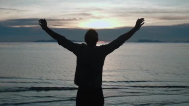 Genç adamın silueti temiz havanın tadını çıkarıyor ve denizde gün batımını seyrediyor. Arkadan bakınca, erkek onun önüne bakıyor. Yüksek kalite 4k görüntü