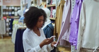 Akıllı telefon kullanan Afro-Amerikalı kadın, moda mağazasında askıda asılı duran yeni kıyafet dokularının bilgilerini kontrol ediyor.