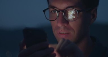Gözlüklü genç adam, akıllı telefondan sayfa uygulaması, sosyal ağa göz at, akıllı telefon ekranı gözlüğe yansıyor. Akşamları açık havada akıllı telefondan internet sosyal medyasında gezinen bir adamın portresi