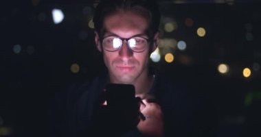 Cep telefonu kullanan genç adamın portresi, uygulama sayfalarını kaydıran sosyal ağlar, yüzüne yansıyan mobil ekran. Gece şehrinin cep telefonu arka planında, gözlüklü bir erkek internete yükleniyor.