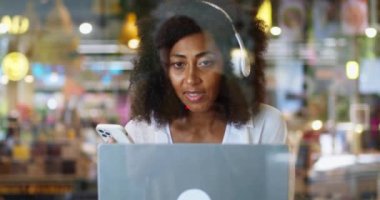 Kulaklıklı Afrikalı Amerikalı bir iş kadını kafede oturmuş laptopla iş birliği yapıyor. Milenyum iş kadını uzaktan kumandası çalışıyor. Telekomünikasyon kavramı. Pencereden yansımayı göster.