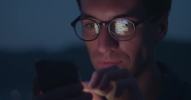 近视男性眼镜滚动页面应用程序在手机上 浏览社交网络 手机屏幕反映在眼镜 男子晚间在户外用手机浏览互联网社交媒体的肖像 — 图库视频影像