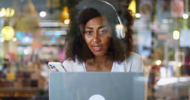 Kulaklıklı Afrikalı Amerikalı Bir Kadını Kafede Oturmuş Laptopla Birliği Yapıyor — Stok video