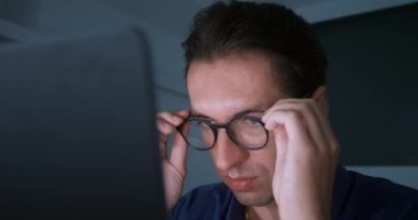 Close-up of pensive overworked businessman working late on laptop in evening home office. Sad businessman takes off glasses and thinks solving business problems, computer monitor reflected in glasses