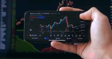 Close up trader analyzing stock market investments on smartphone with chart of stocks or cryptocurrencies on background. Person trader holding using app smartphone for trading stock exchange.