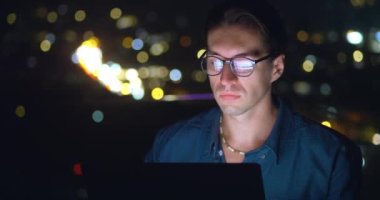 Portrait of a male student working remotely on a laptop at home on a balcony against the backdrop of a night city. A student with glasses is studying online using a laptop computer