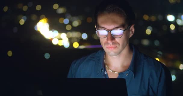 ポートレートの男性のフリーランスは 夜の街を背景にノートパソコンでリモートで動作します ガラスの男性の千年紀30夜にラップトップコンピュータの屋外を使用して コンピュータモニタはガラスに反映されます — ストック動画