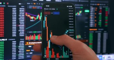 Crypto investor using mobile phone app for buying cryptocurrency in front of stock market financial screen, close up. Trader investor holding mobile phone analyzing financial crypto stock market price