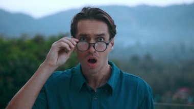 Frightened man looks at camera and takes off glasses in surprise, covering his mouth with hand in fear, in evening on backdrop of nature and mountains. Close-up frightened male with glasses outdoors.