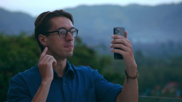 ミレニアル世代の男性は ビデオ通話にスマートフォンを使用し 自然を背景に友人と話をします 眼鏡と青いシャツの若い男の観光客は 熱帯国の休暇中にビデオ通話を介してスマートフォンで話す — ストック動画