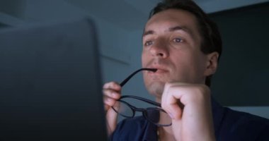 Close-up of pensive man entrepreneur working on laptop in evening at home office. Man takes off glasses and thinks about new ideas for solving business problems, computer monitor reflected in glasses