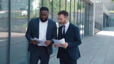 Two male top managers in suits discussing work project standing outside office building. Diverse male top managers hold paper documents in hands and talk about business, during lunch break near office
