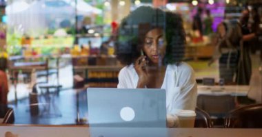 Confident multi-ethnic woman, manager or sales representative making call on smartphone, drinking takeaway hot drink from paper cup, working on laptop computer indoors. Working from coffee shop.
