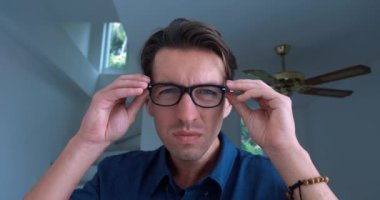 Portrait of a young man, 30 years old, in a blue shirt, puts on glasses to see better and smiling against the backdrop of an indoor home office. Correction of vision with glasses.