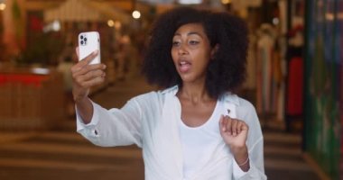 Slow motion: dark skinned lady enjoys distance communication, uses smartphone and mobile apps,makes a video call, talks via video link, has pleasant smile, dressed in white clothes, in store interior