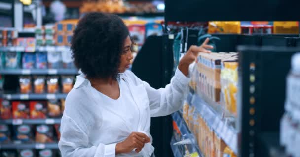 アフリカ系アメリカ人の若い女性の買い物客は 白いカジュアルなシャツと青いデニムジーンズで 食料品店でシリアルを買い物している間 ラベルの内容を注意深く読む — ストック動画